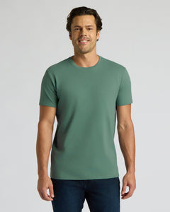 True ClassicA man wearing a solid sage green crew-neck t-shirt and dark blue jeans, standing against a light gray studio background.