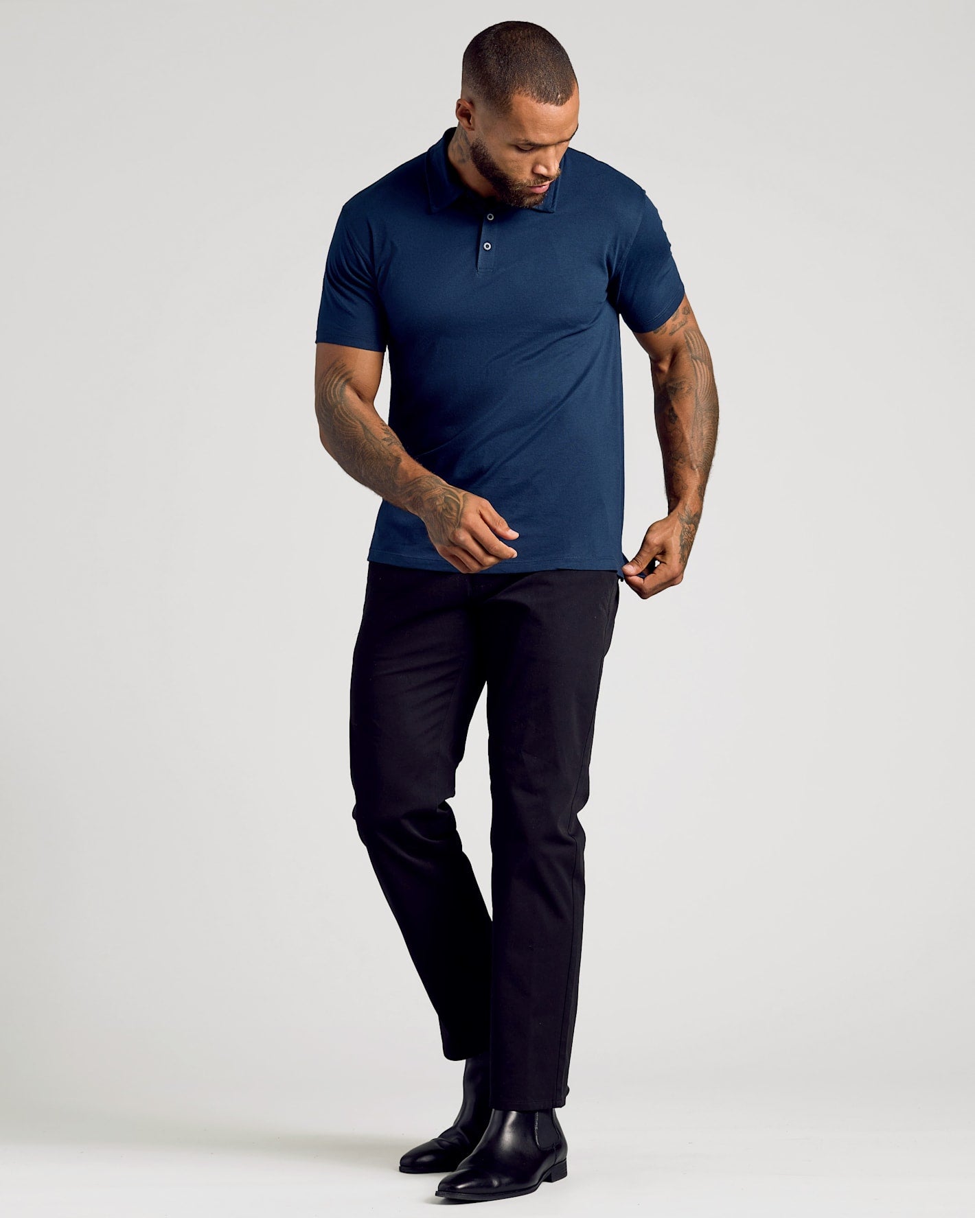 A man modeling a navy blue short-sleeve polo shirt with black trousers and black leather shoes, standing against a light gray studio background.