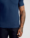 A close-up of a person wearing a solid navy blue short-sleeve t-shirt and dark pants, showing the side of the torso and tattooed arm against a light gray background.
