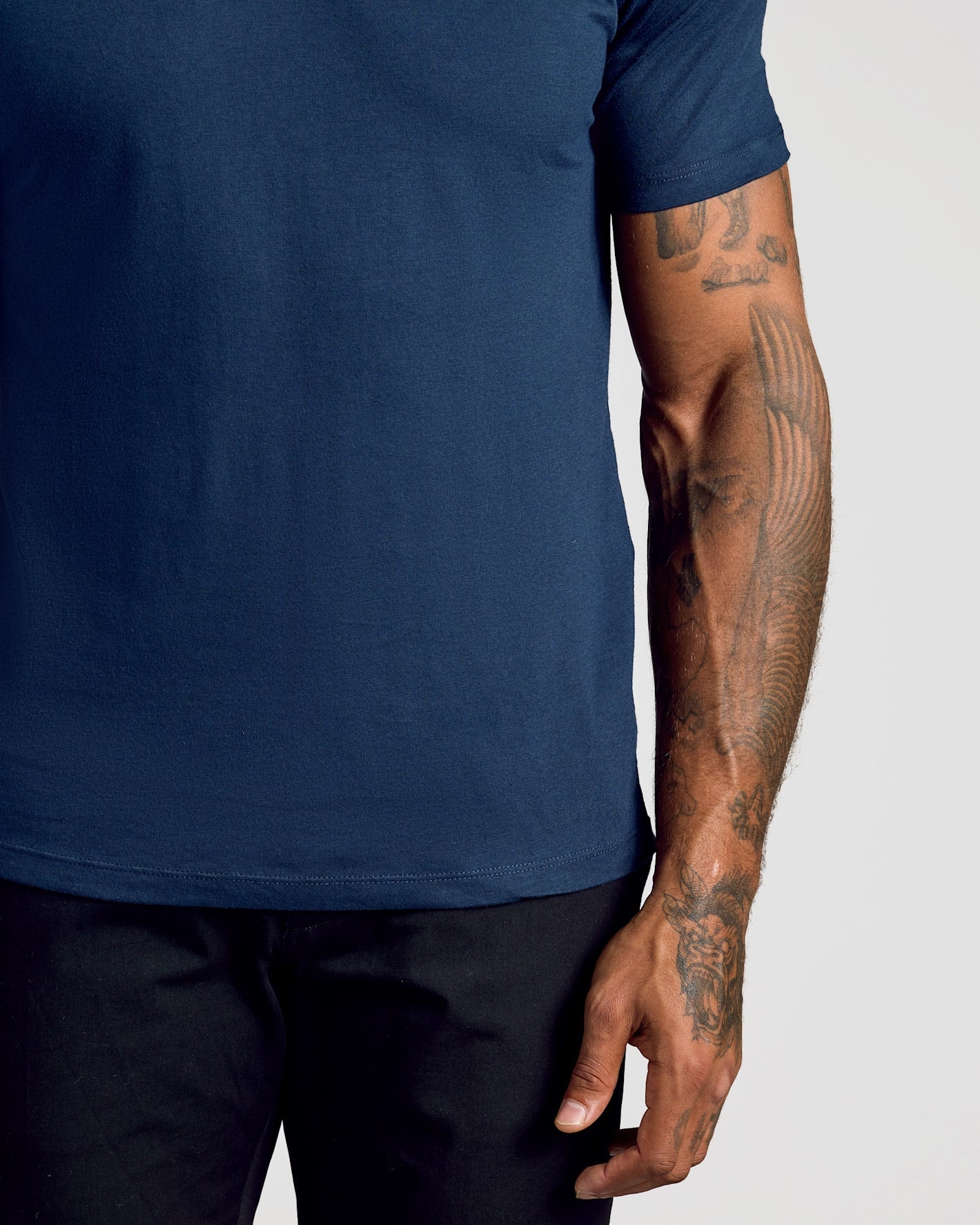 A close-up of a person wearing a solid navy blue short-sleeve t-shirt and dark pants, showing the side of the torso and tattooed arm against a light gray background.