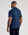 A man viewed from the back wearing a navy blue short-sleeve polo shirt and dark trousers, standing against a plain light gray studio background.