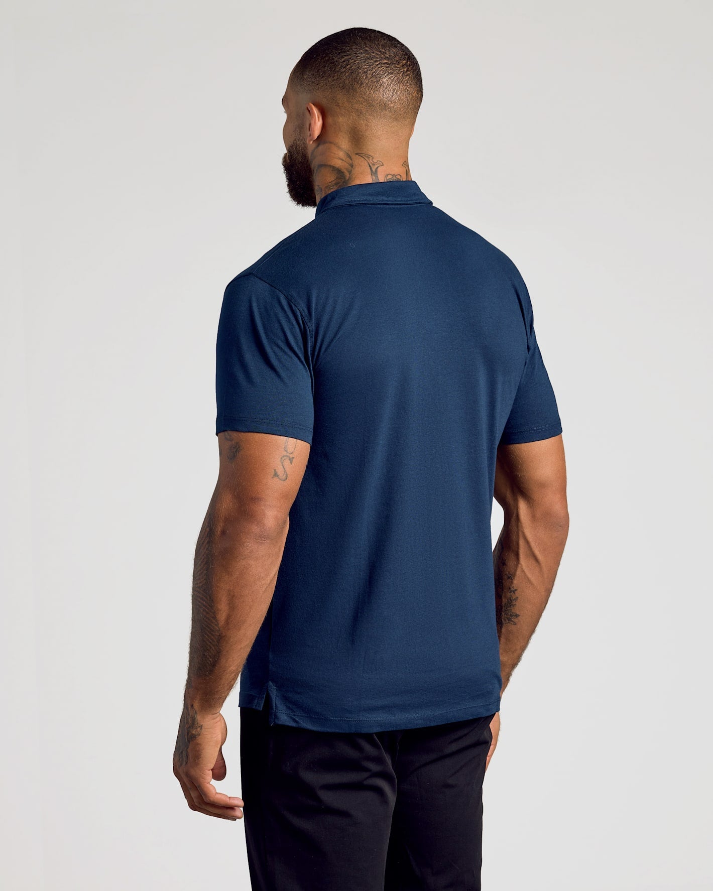 A man viewed from the back wearing a navy blue short-sleeve polo shirt and dark trousers, standing against a plain light gray studio background.
