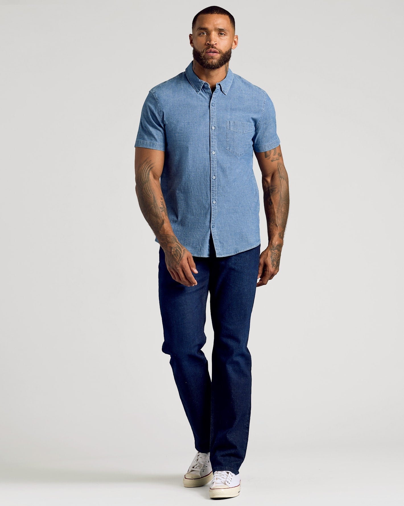 A man modeling a light blue short-sleeve button-down shirt with dark blue jeans and white sneakers, standing against a light gray studio background.