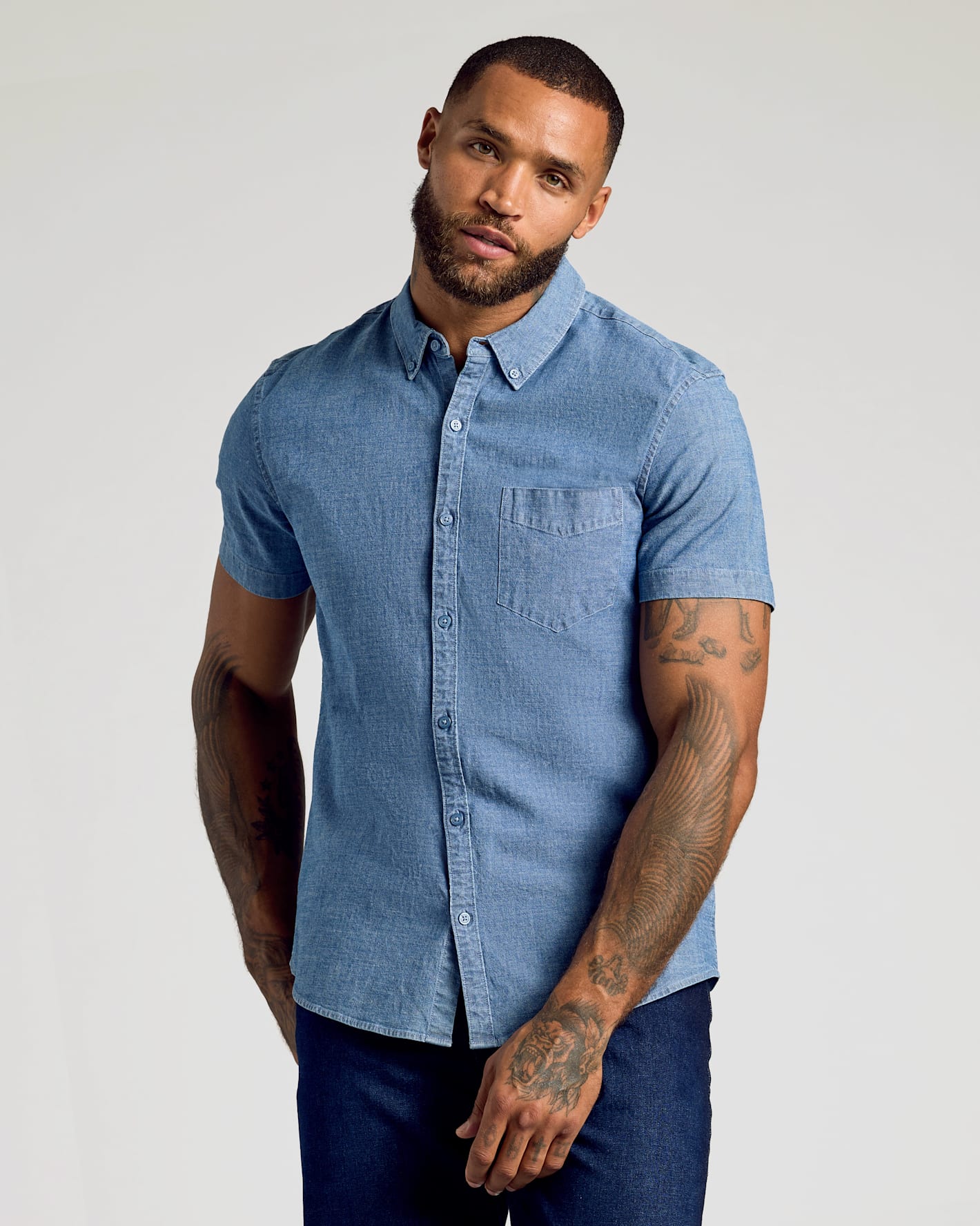 A man wearing a light blue short-sleeve button-down shirt and dark blue jeans, standing against a light gray studio background.