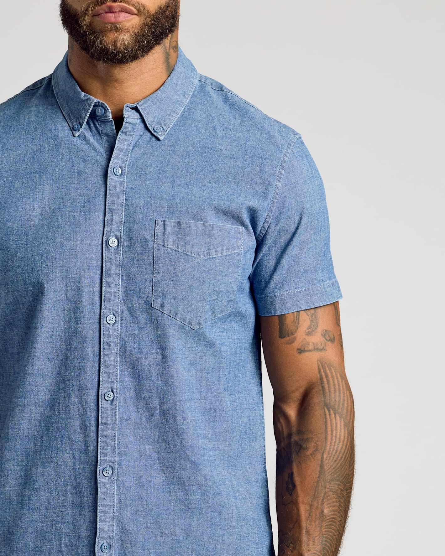 A close-up of a man wearing a light blue chambray short-sleeve button-down shirt, showing the chest pocket and fabric texture against a neutral light gray background.