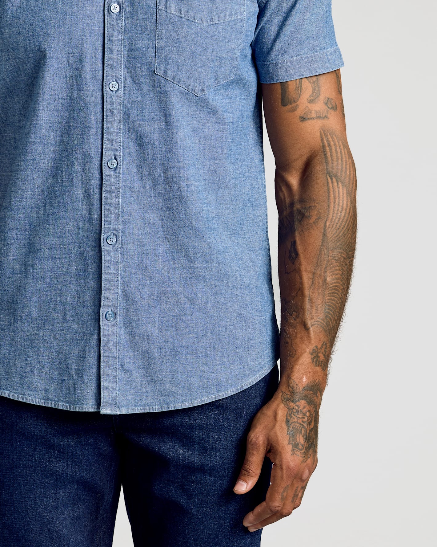 Close-up of a man wearing a light blue chambray short-sleeve button-up shirt and dark blue jeans, showing his tattooed arm against a light gray background.