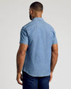 A man viewed from the back wearing a light blue short-sleeve chambray button-down shirt and dark blue jeans, set against a plain light gray studio background.