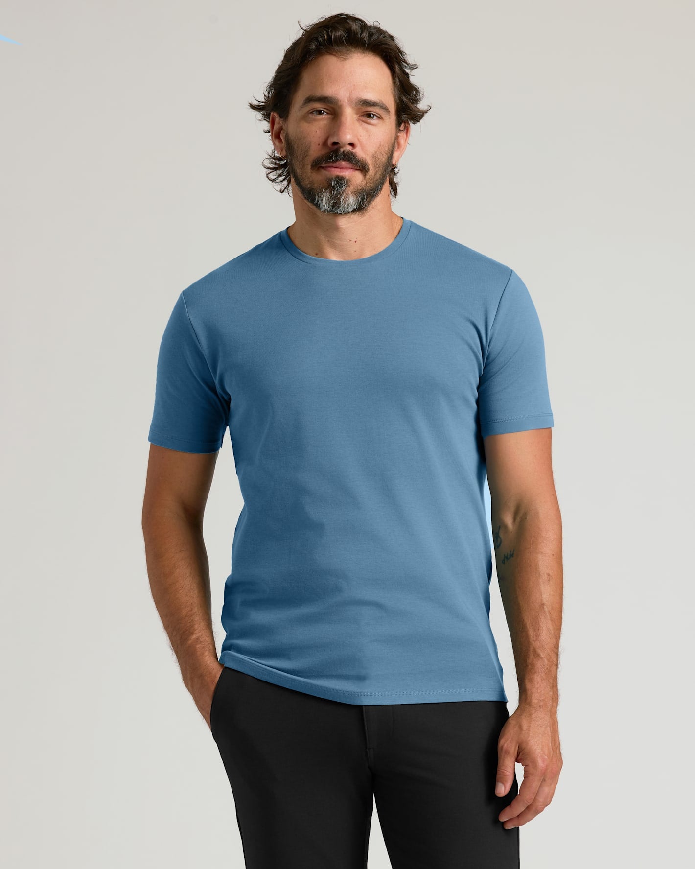 A man wearing a solid medium blue crew-neck t-shirt and black pants, standing against a light gray studio background.