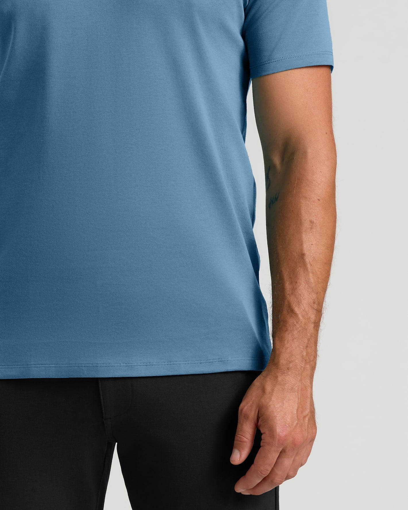A close-up of a man wearing a short-sleeve crew-neck t-shirt in a medium blue color, paired with black pants, against a light gray background.