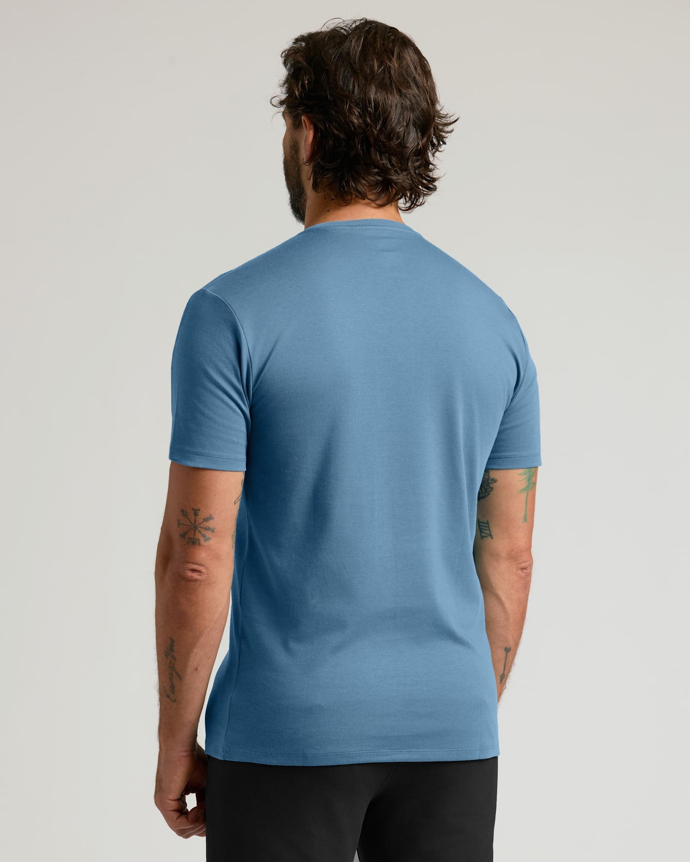 A man viewed from behind wearing a short-sleeve crew-neck t-shirt in a medium blue color, paired with black pants, against a light gray studio background.