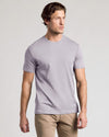 A man wearing a light lavender crew-neck t-shirt and khaki pants, standing against a neutral light gray background.