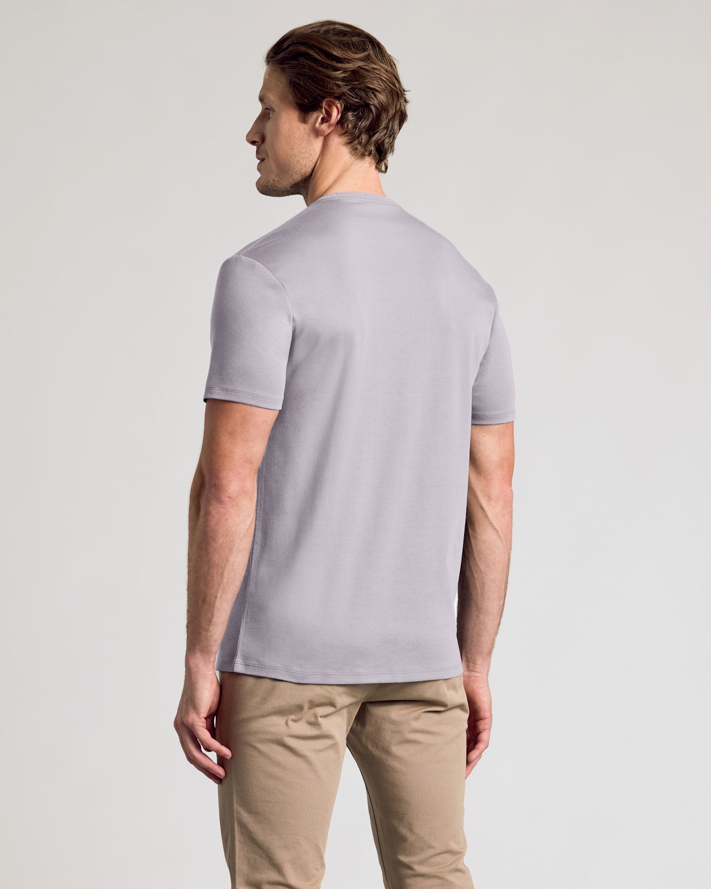 A man wearing a light gray short-sleeve t-shirt and khaki pants, shown from a rear three-quarter view against a plain light gray studio background.