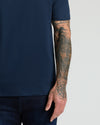 Close-up of a man's arm with detailed tattoos wearing a Navy Pima Cotton Blend Crew shirt paired with dark jeans against a plain light background.