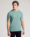 A man wearing a short-sleeve crewneck t-shirt in a muted sage green color, paired with black pants, standing against a light gray studio background.
