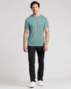 A man modeling a short-sleeve crewneck t-shirt in a muted teal green color, paired with black pants and white sneakers, against a plain light gray studio background.