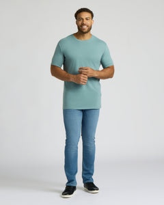 True ClassicA man modeling a short-sleeve crewneck t-shirt in a dusty teal color, paired with blue jeans and black slip-on shoes, standing against a light gray studio background.