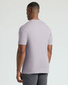 A man viewed from the back wearing a light lavender short-sleeve crewneck t-shirt and gray pants, set against a plain light gray studio background.