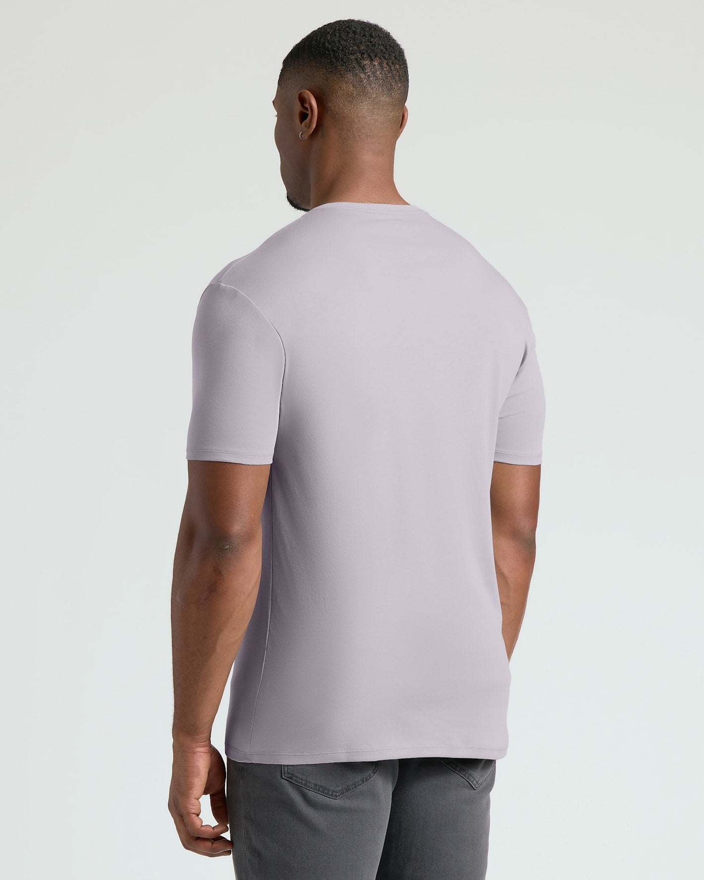 A man viewed from the back wearing a light lavender short-sleeve crewneck t-shirt and gray pants, set against a plain light gray studio background.