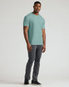 A man modeling a short-sleeve crewneck t-shirt in a muted sage green color, paired with dark gray pants and black sneakers, against a plain light gray studio background.