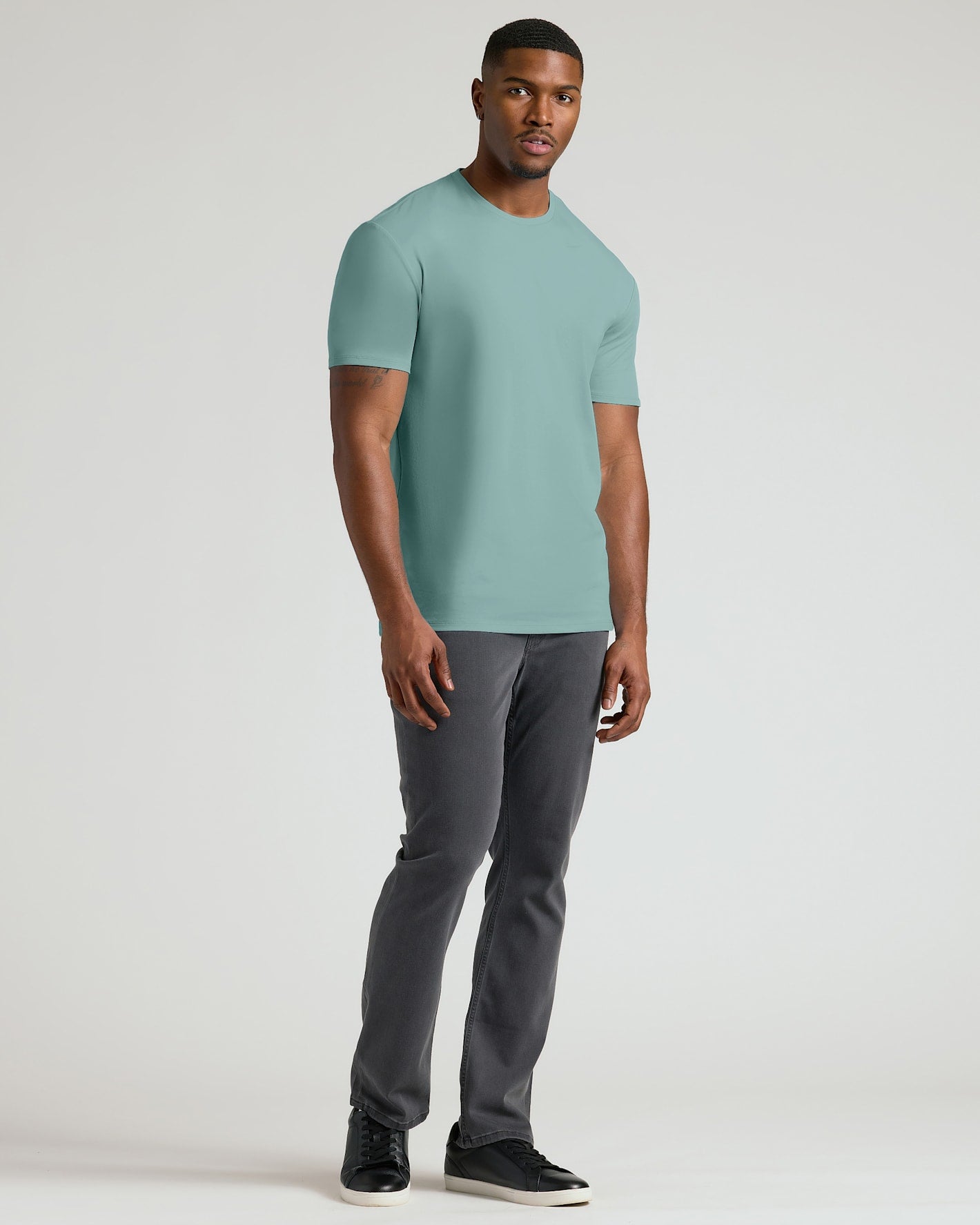 A man modeling a short-sleeve crewneck t-shirt in a muted sage green color, paired with dark gray pants and black sneakers, against a plain light gray studio background.