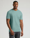 A man wearing a solid sage green crew-neck t-shirt and dark gray pants, standing against a light gray studio background.