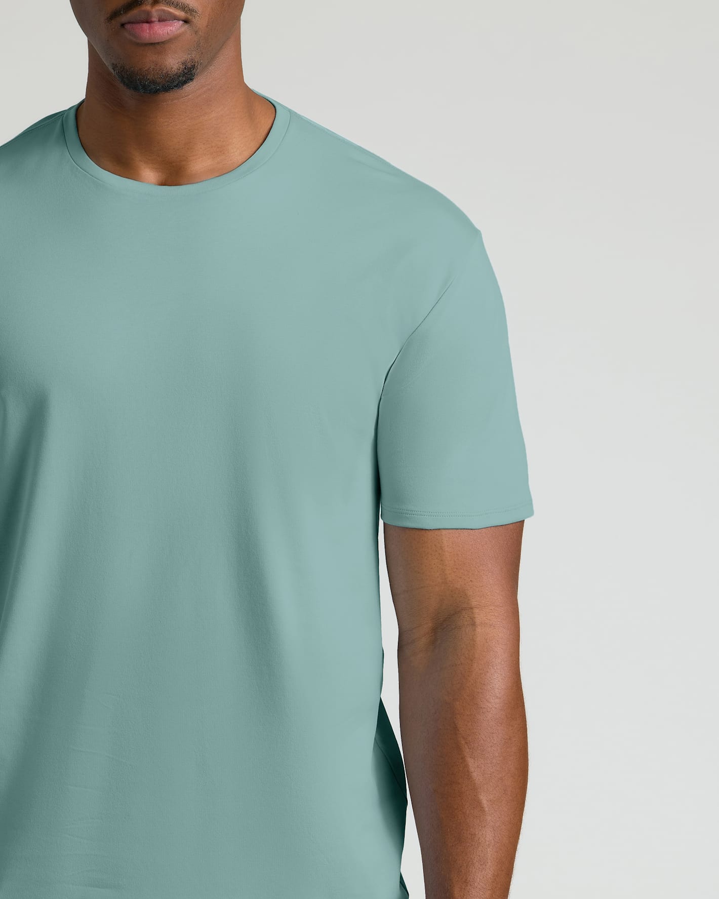 A close-up of a man wearing a solid sage green crew-neck t-shirt against a light gray studio background.