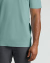 A close-up of a person wearing a short-sleeve crew-neck t-shirt in a muted sage green color, paired with dark gray jeans, against a light gray background.