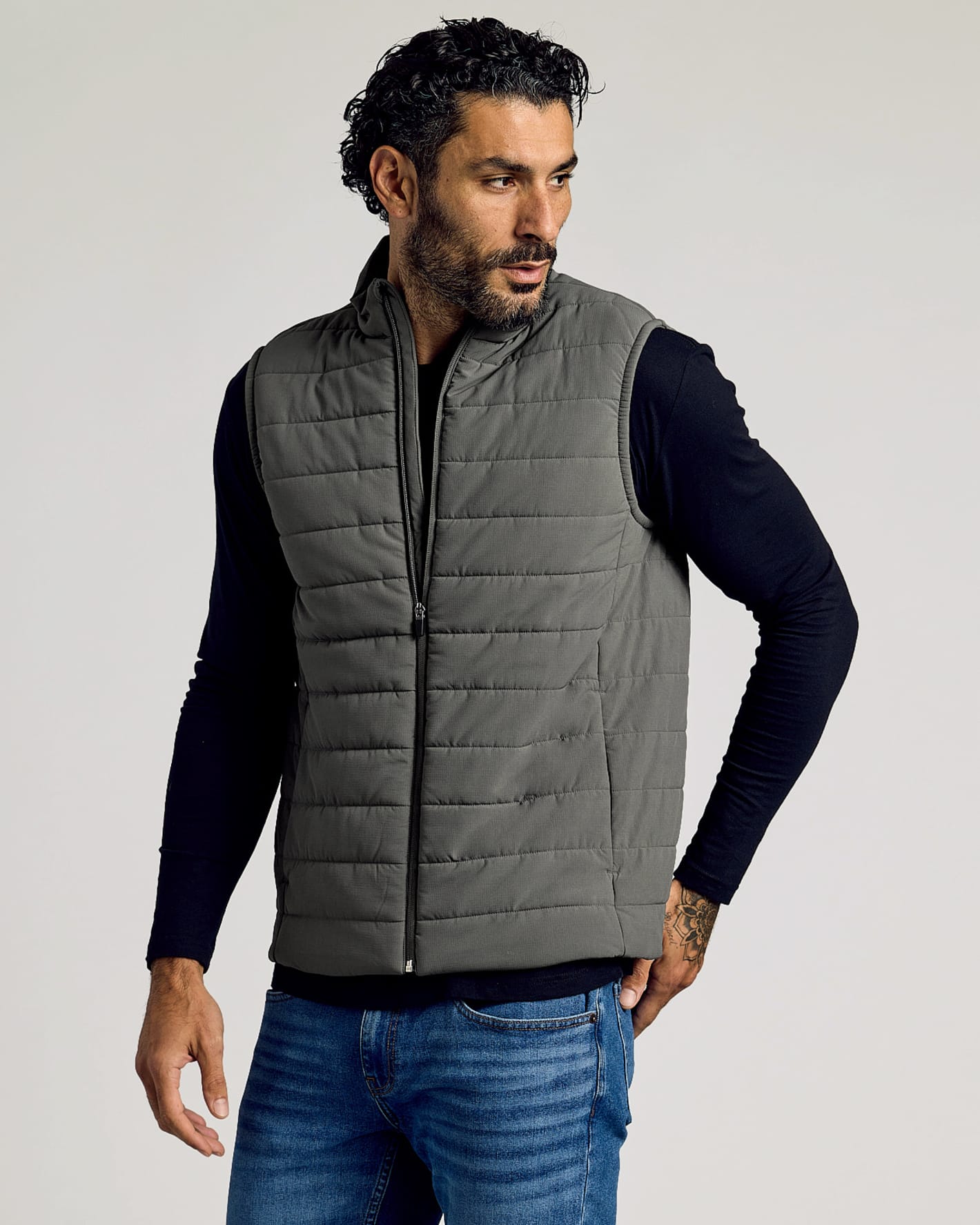 Man wearing a gray Carbon Puffer Vest over a black long-sleeve shirt and blue jeans, showcasing a casual and comfortable men's basics look.
