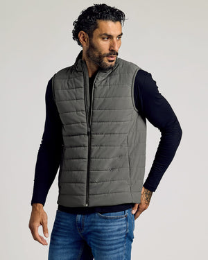 Man wearing a gray Carbon Puffer Vest over a black long-sleeve shirt and blue jeans, showcasing a casual and comfortable men's basics look.