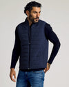 Man wearing a navy puffer vest over a black long-sleeve shirt and blue jeans, posing with one hand in pocket and looking to the side in a studio setting