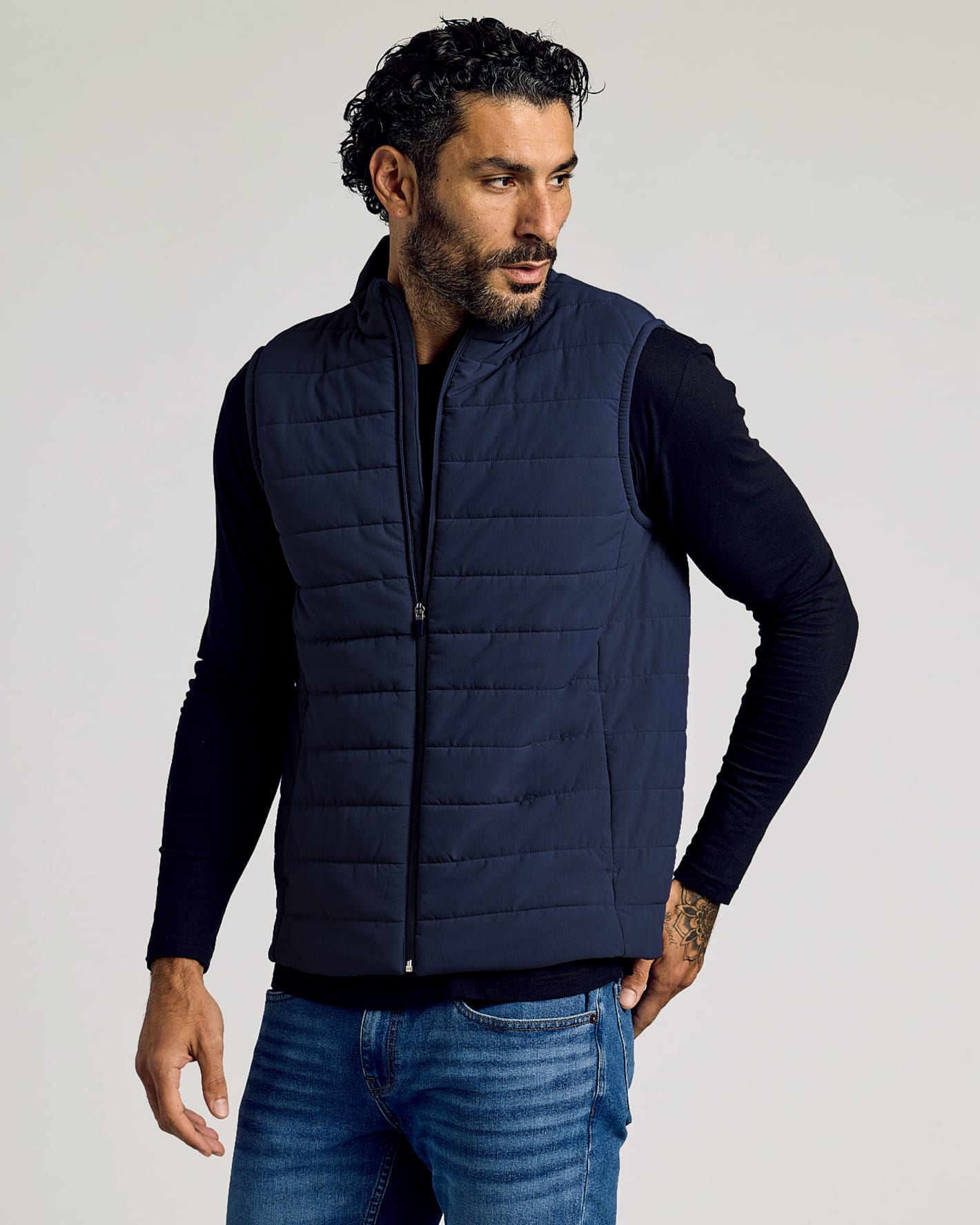Man wearing a navy puffer vest over a black long-sleeve shirt and blue jeans, posing with one hand in pocket and looking to the side in a studio setting