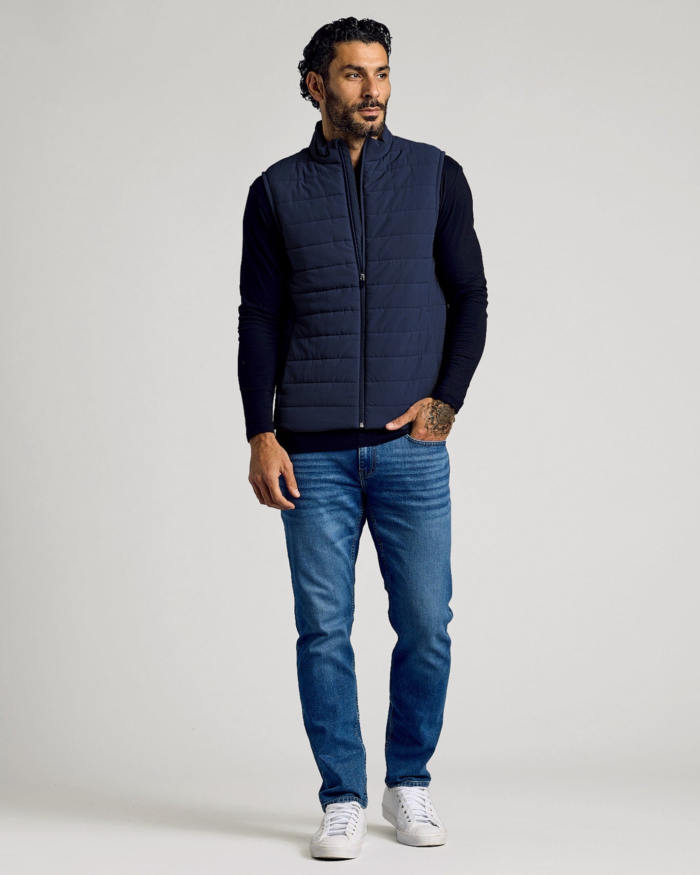 Man wearing a Navy Puffer Vest over a black long sleeve shirt paired with blue jeans and white sneakers, posing against a plain background in a casual style.