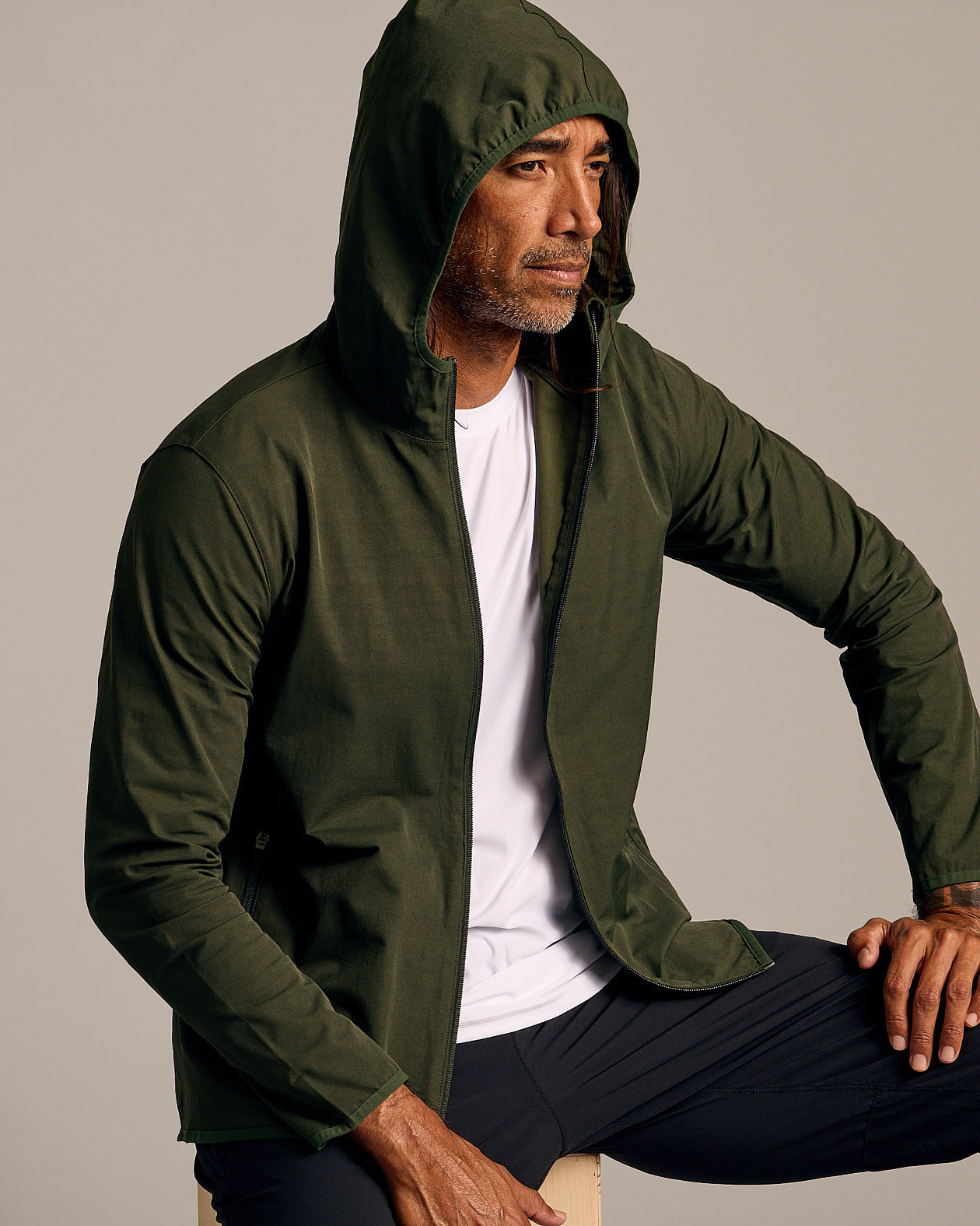 Olive Tech Windbreaker on seated man, hood up, casual pose.