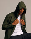 Man wearing Olive Tech Windbreaker with hood up, holding jacket edges.