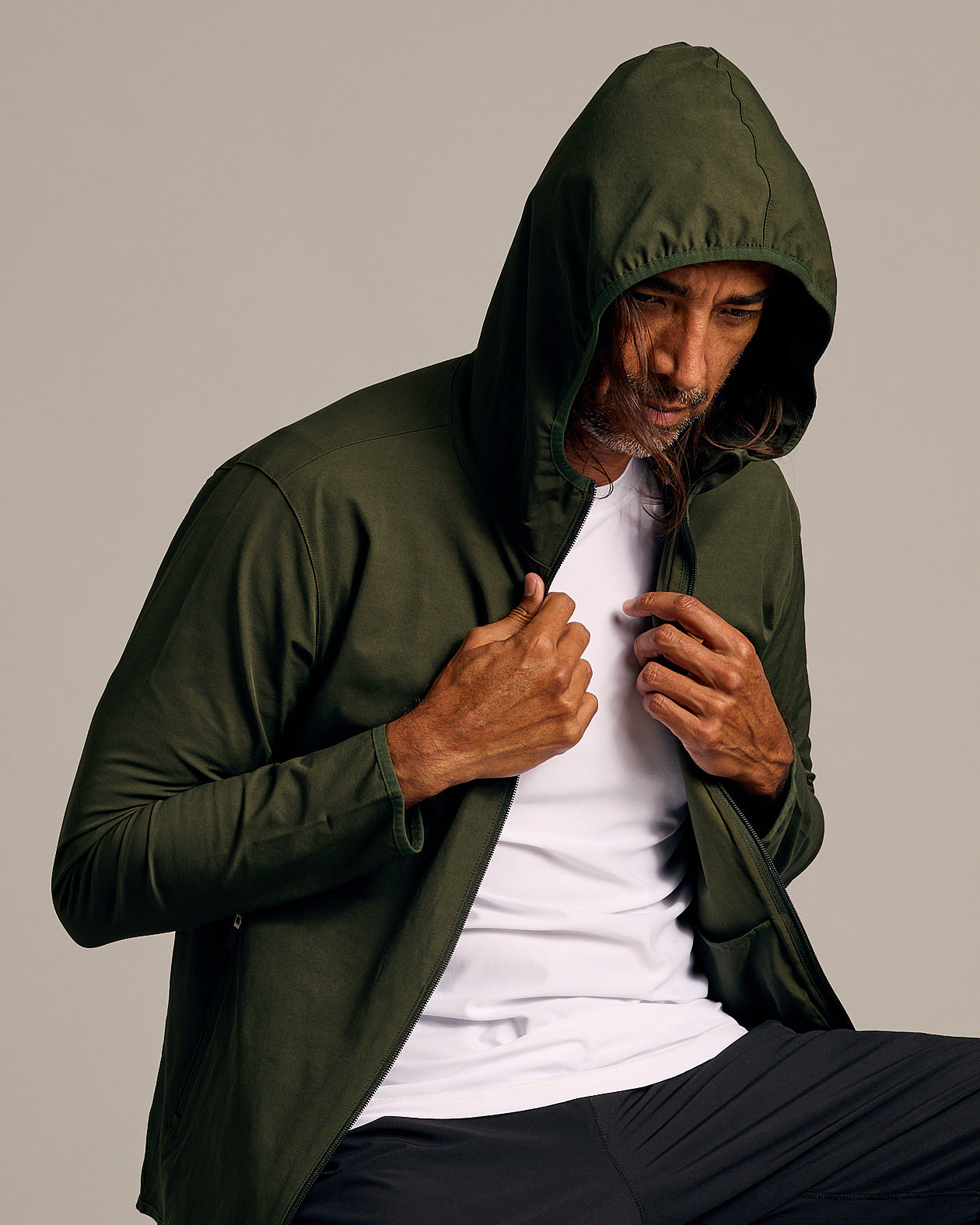 Man wearing Olive Tech Windbreaker with hood up, holding jacket edges.