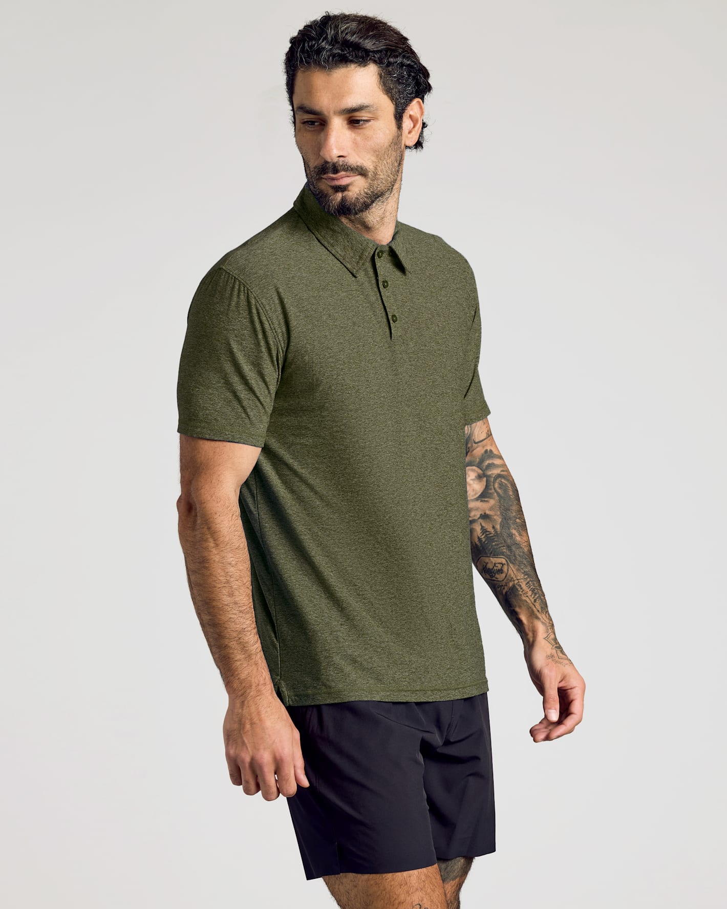 Man wearing a Dark Heather Pear Active Polo 2.0 in olive green, paired with black shorts, standing against a light background in a casual pose.