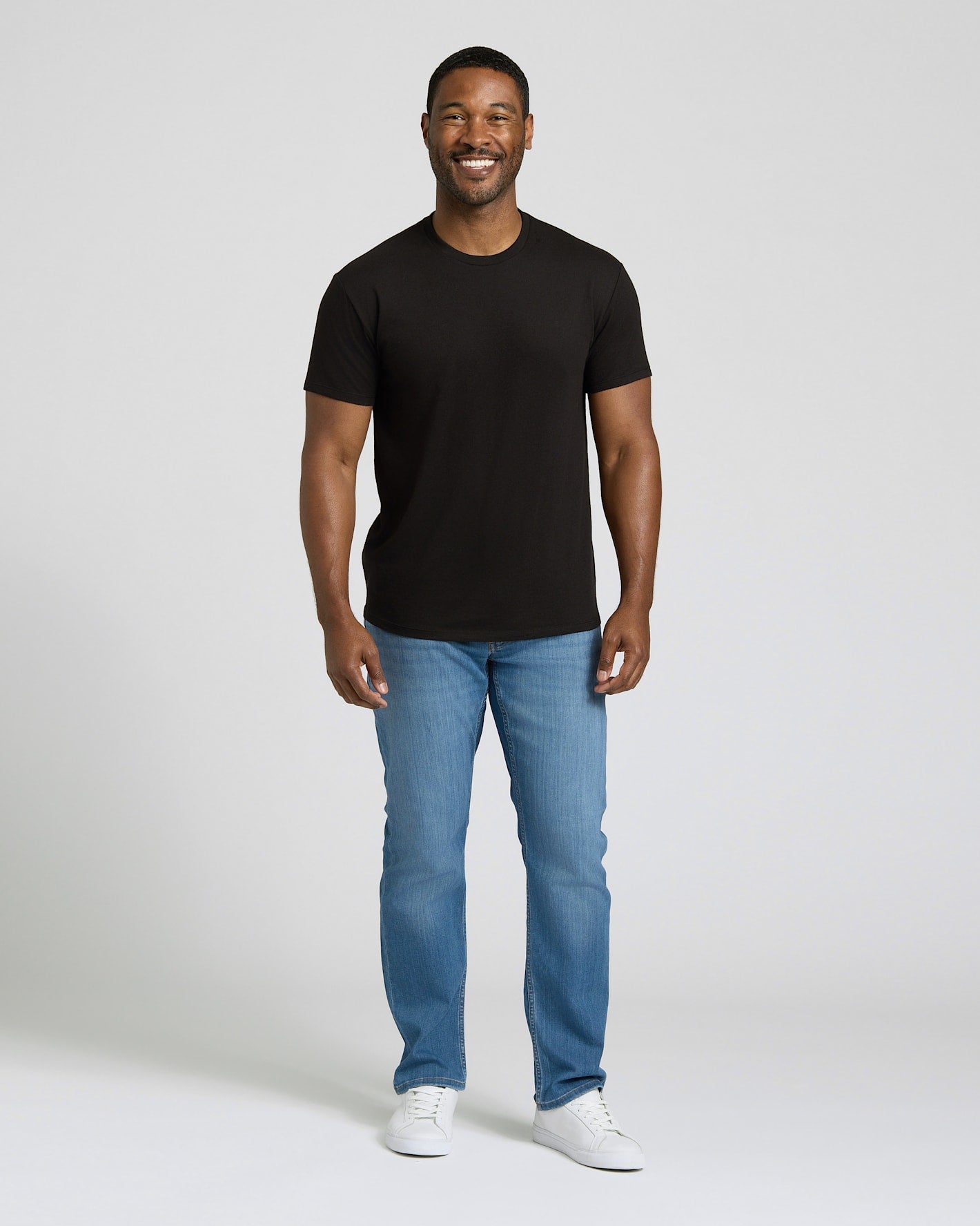 Man wearing a black t-shirt, blue jeans, and white sneakers on a plain background. MODEL_INFO: Model is 5'8" wearing size M short