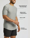 A man wearing a light heather gray short-sleeve athletic t-shirt with black shorts, shown against a light gray background with text callouts highlighting features like sweat-wicking, odor-blocking, stretch, built-in SPF, and curved hem.