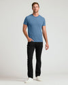 A man modeling a short-sleeve crewneck t-shirt in a medium blue color, paired with black pants and white sneakers, standing against a light gray studio background.