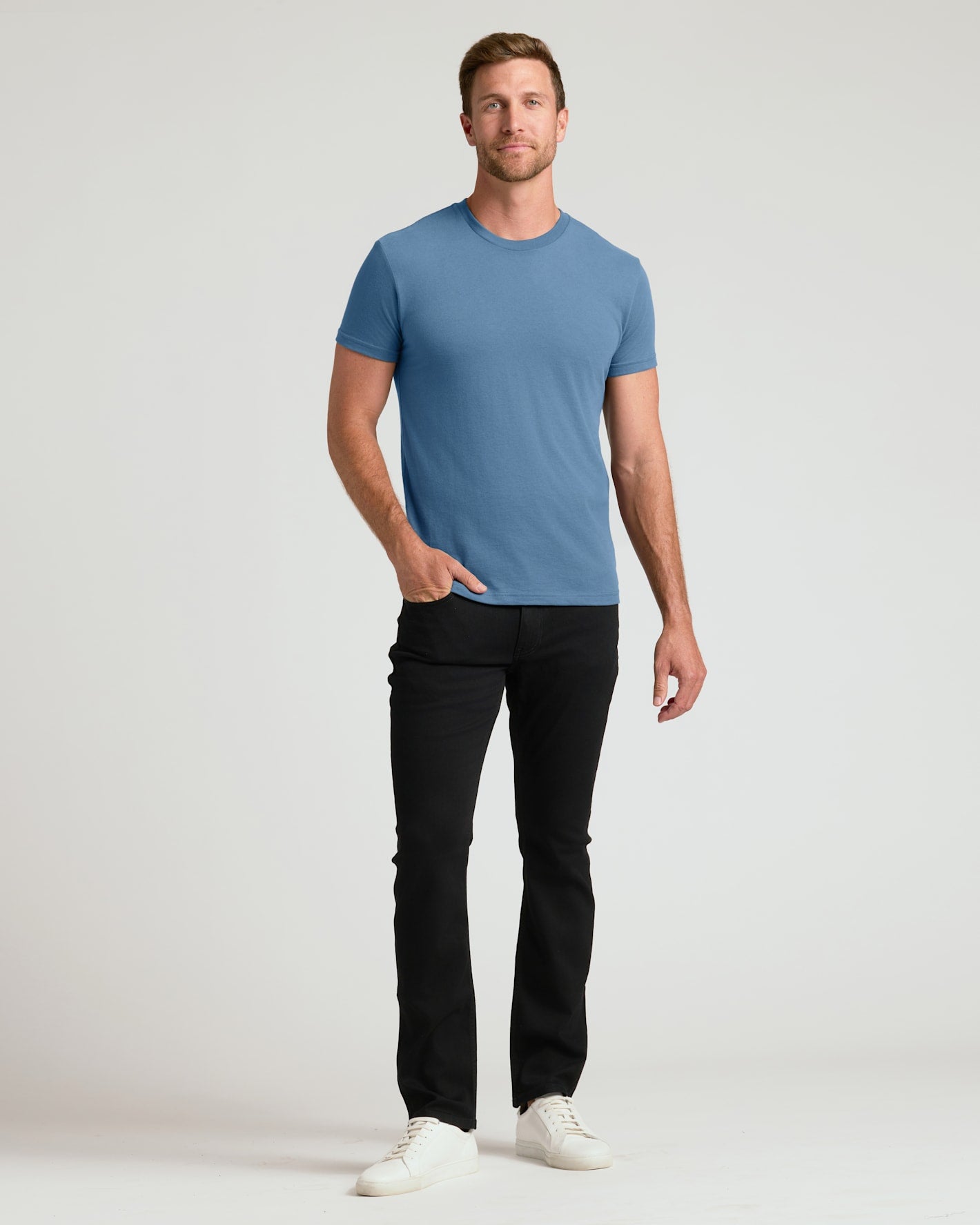 A man modeling a short-sleeve crewneck t-shirt in a medium blue color, paired with black pants and white sneakers, standing against a light gray studio background.