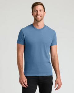True ClassicA smiling man wearing a solid medium blue crew-neck t-shirt and black pants, standing against a light gray studio background.