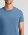 A man wearing a solid medium blue crew-neck t-shirt, shown from the chin down against a light gray studio background.
