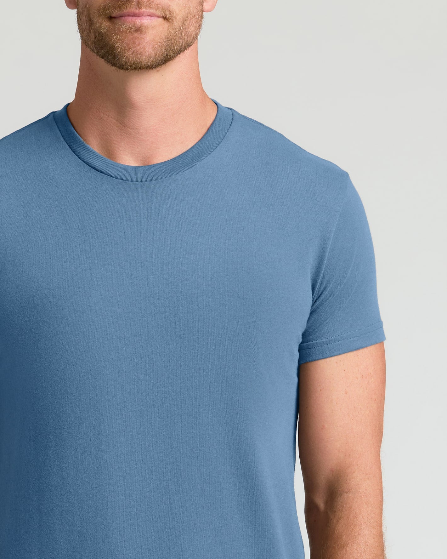 A man wearing a solid medium blue crew-neck t-shirt, shown from the chin down against a light gray studio background.