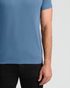 A close-up of a man wearing a solid medium blue short-sleeve t-shirt and black pants, cropped from the chest down against a light gray background.