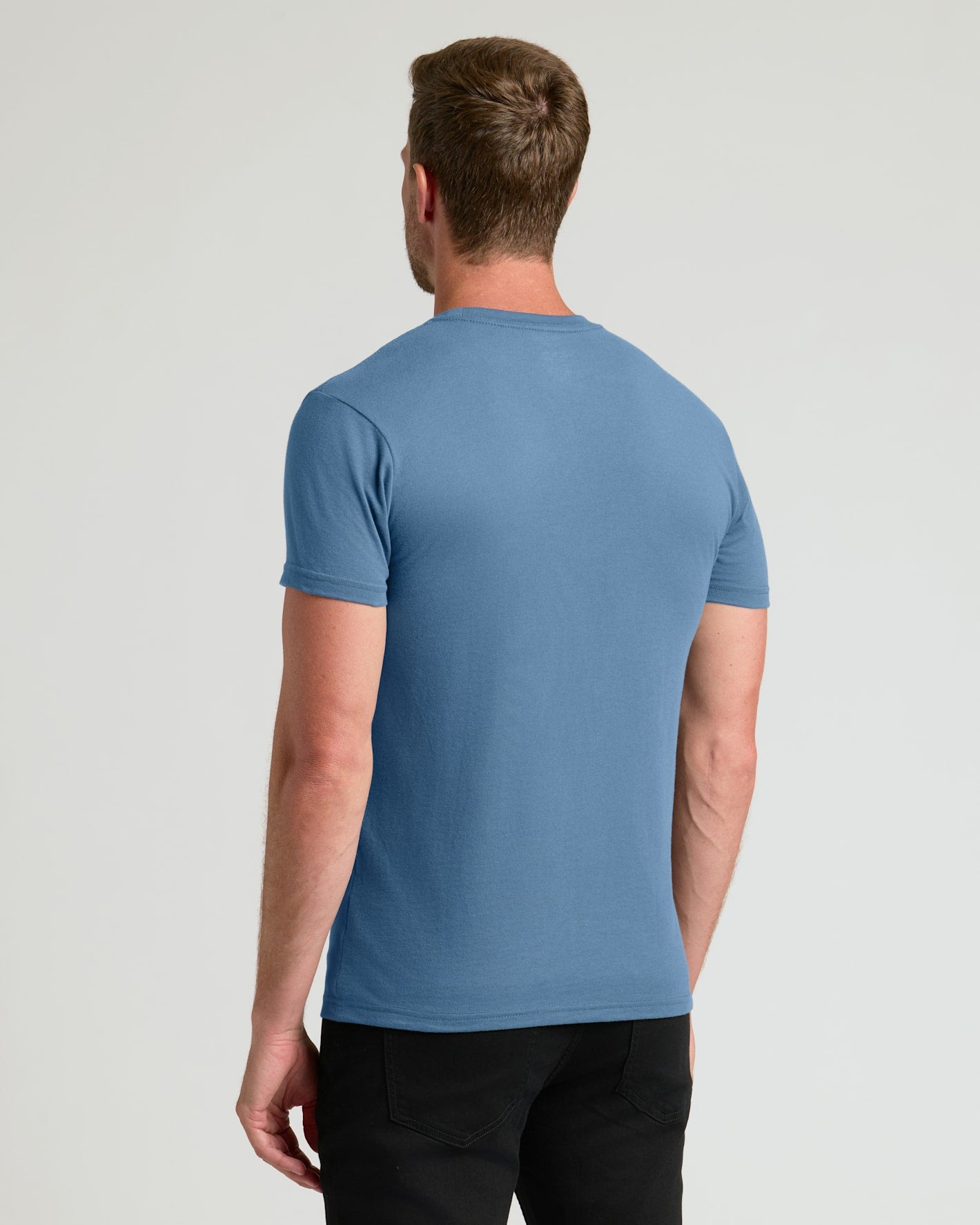 A man viewed from the back wearing a solid medium blue short-sleeve crewneck t-shirt and black pants, set against a plain light gray studio background.