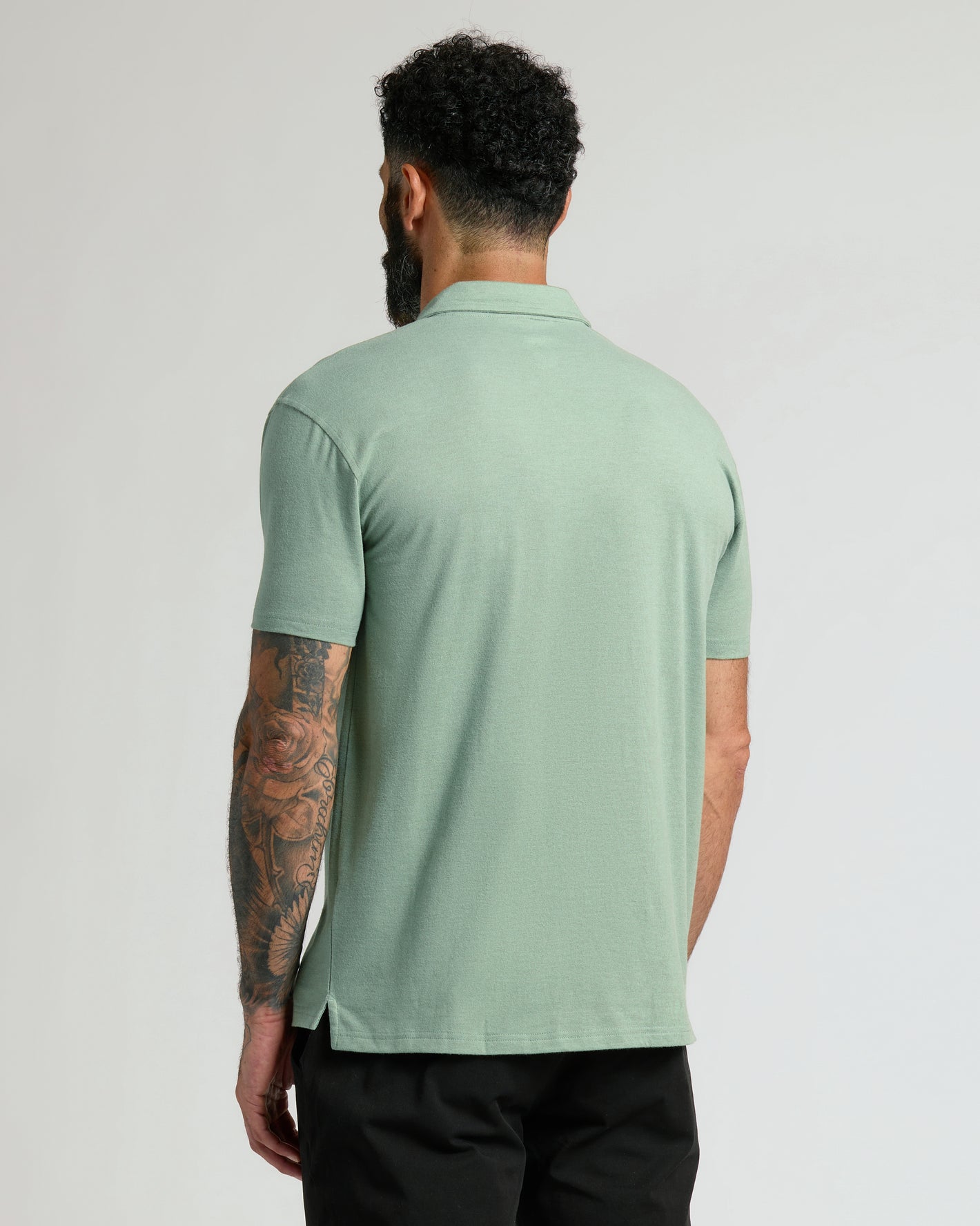 Heather Slate Green Classic Polo, tattooed arm, black pants, back view.