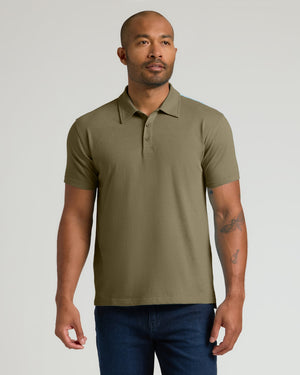 A man wearing a short-sleeve polo shirt in olive green, paired with dark blue jeans, standing against a light gray studio background.