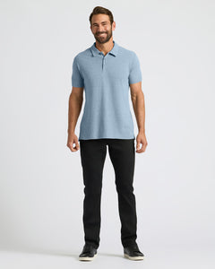 True ClassicA man modeling a light blue short-sleeve polo shirt with black pants and black sneakers, standing against a plain light gray studio background.
