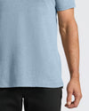 Close-up of a person wearing a light heather blue short-sleeve t-shirt and black pants against a plain light gray background.