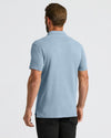 A man viewed from the back wearing a light heather blue short-sleeve polo shirt and black pants, against a plain light gray studio background.